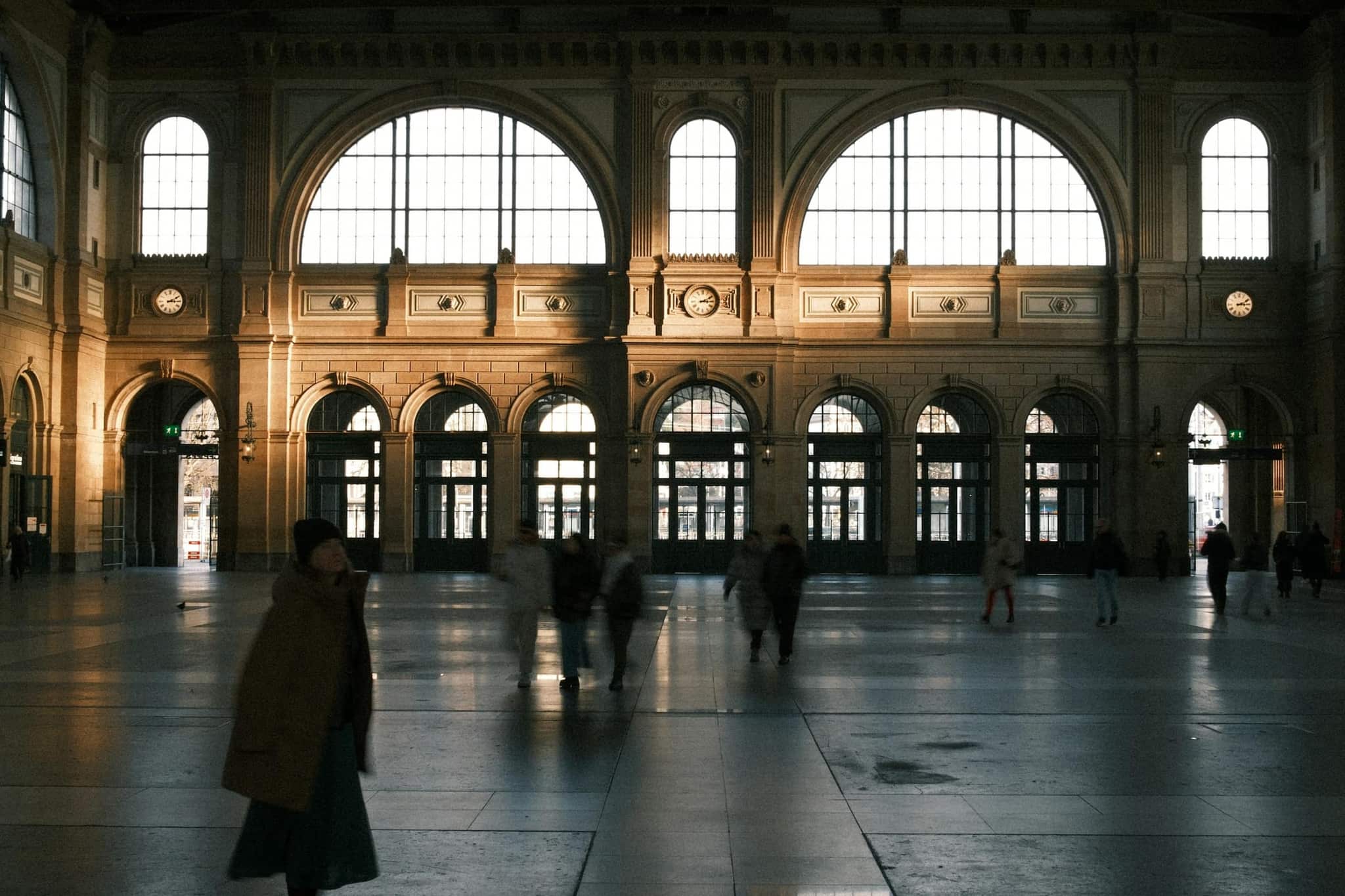 The main concourse hall of Zürich's central railway station (Zürich Hauptbahnhof) at golden hour — a lone traveller in a winter coat crossing the floor toward the tall arched windows, other passengers in soft motion blur.