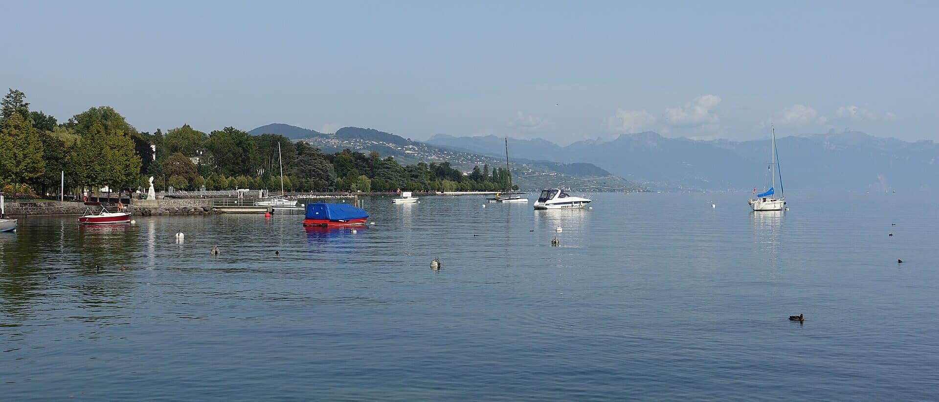 Le lac Léman à Ouchy, Lausanne — la promenade bordée d'arbres et les Alpes dans la brume au-dessus du lac.